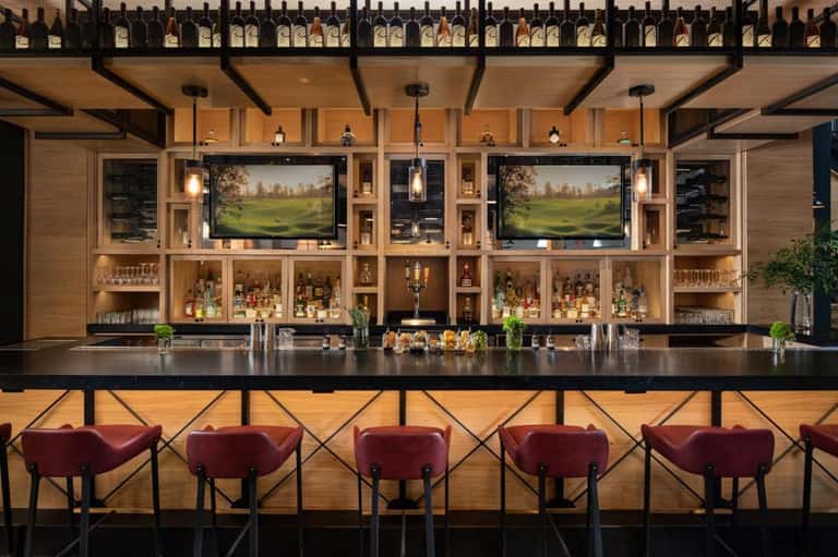 Upscale bar interior with dual TV screens, liquor shelves, and burgundy bar seating