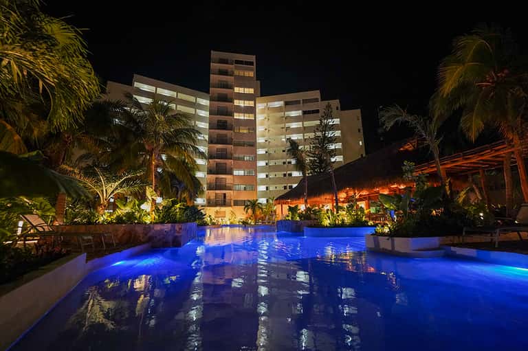 Illuminated resort pool at night with blue lighting, palm trees, and multi-story hotel building in background