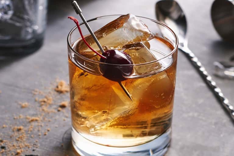 Cocktail with amber liquid, ice, and cherry garnish with red stirrer on gray surface