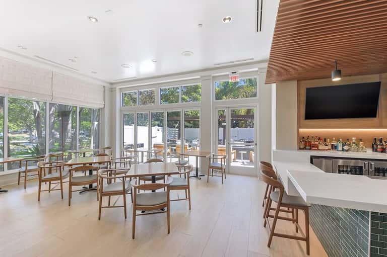 Bright cafe space with wooden chairs, white bar counter, large windows, and outdoor seating visible