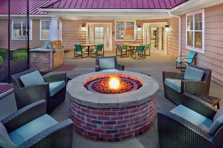 Outdoor fire pit patio with woven lounge chairs, brick surround, purple metal roof house, and landscaping