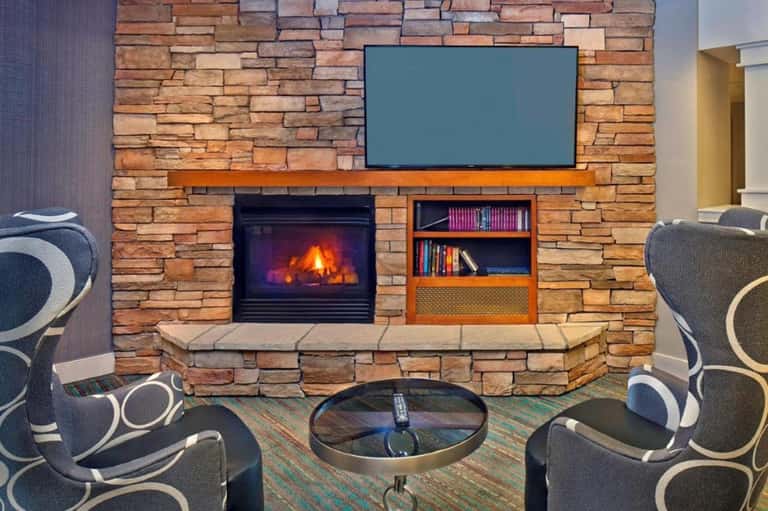 Stone fireplace with lit fire, TV above mantel, and patterned armchairs in modern lounge setting