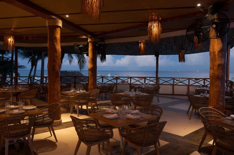 Beachfront restaurant with wooden columns, ocean view, wicker chairs, and sunset lighting