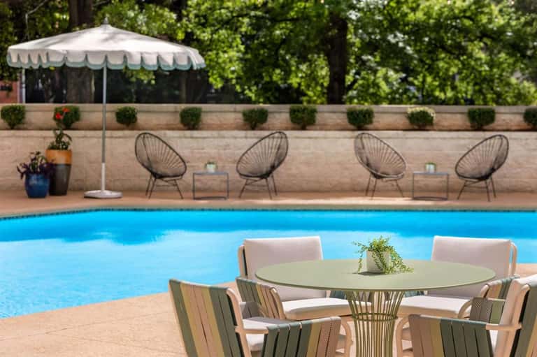 Poolside lounge area with round tables, wire chairs, white umbrella, and stone wall backdrop