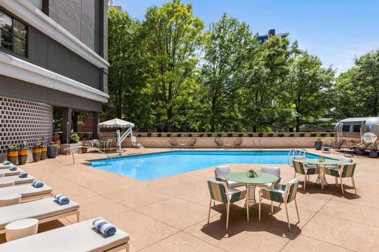 Resort pool deck with lounge chairs, green seating areas, and modern building with tree-lined backdrop