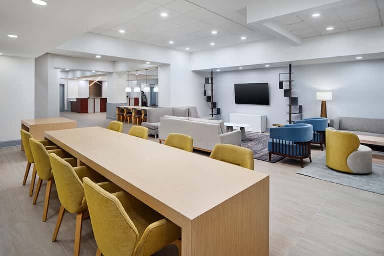 Modern hotel gathering space with dining table, yellow chairs, lounge seating, and reception desk