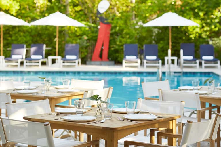 Poolside dining area with wooden tables, white chairs, umbrellas, and lounge seating