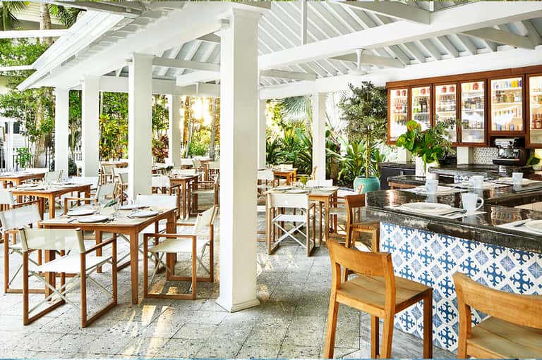 Covered dining patio with white columns, wooden tables, blue accents, and tropical plants