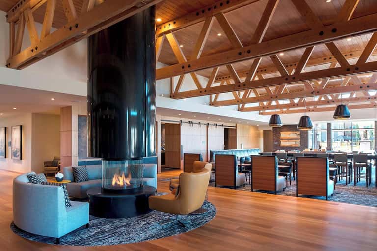 Modern hotel lobby with central fireplace, exposed wooden beam ceiling, blue seating areas, and bar in background