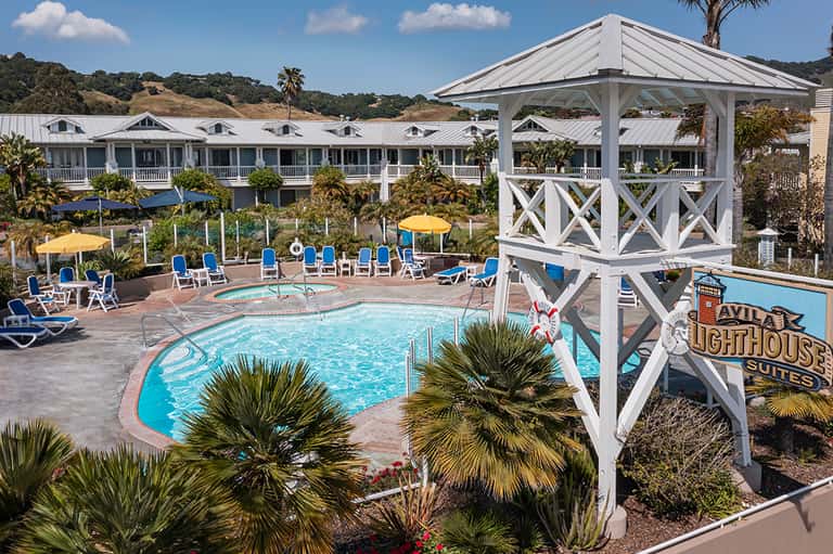 Resort pool area with blue loungers, yellow umbrellas, white gazebo, palm trees, and hillside accommodations