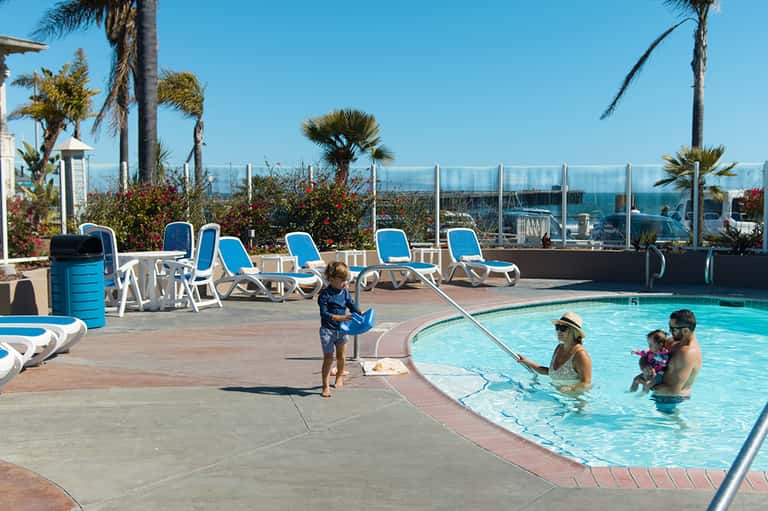 Rooftop pool deck with families swimming, lounge chairs, and ocean views behind glass railing