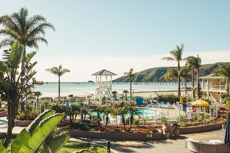 Beach resort with palm trees, pier, pool area, and sandy shoreline with coastal gazebo in summer sunlight