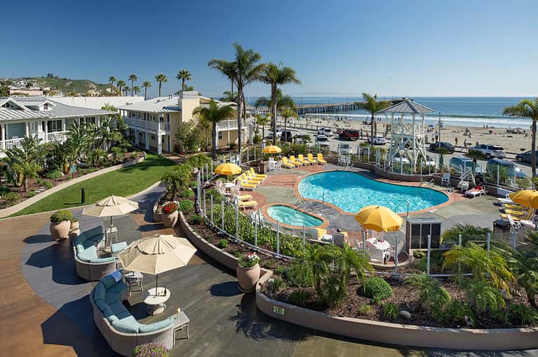 Beachfront resort with curved swimming pool, yellow umbrellas, ocean view, and beach pier in background