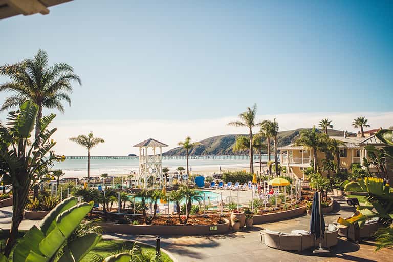Beachfront resort with pool, palm trees, sandy beach, pier, and ocean view with hillside backdrop