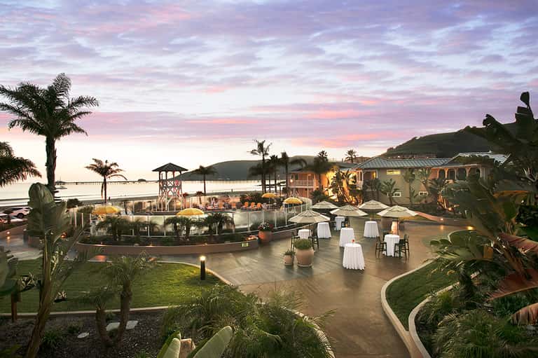 Sunset beach resort with illuminated buildings, white-draped tables, palm trees, and waterfront views