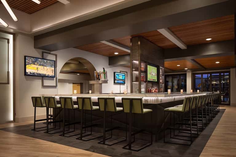 Modern hotel bar with dark wood ceiling beams, olive-green bar stools, multiple TV screens, and contemporary lighting