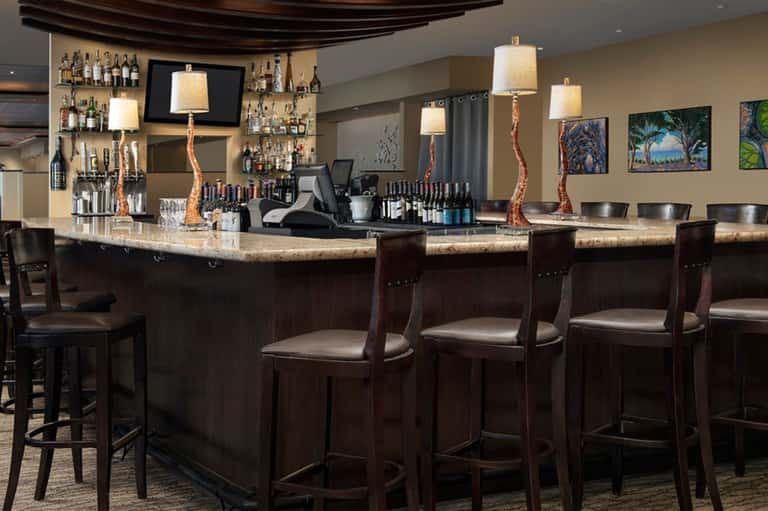 Hotel bar lounge with dark wood counter, bar stools, bottle shelves, and hanging pendant lights