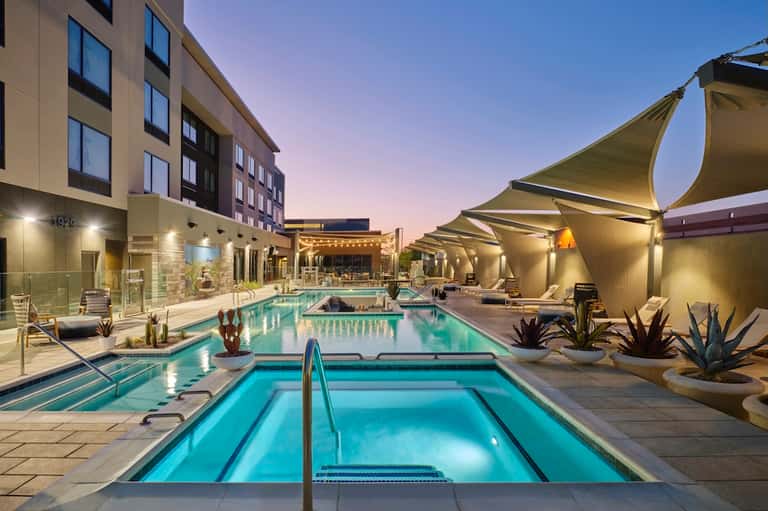 Modern resort pool at twilight with sail shade structures and hotel building illuminated in background