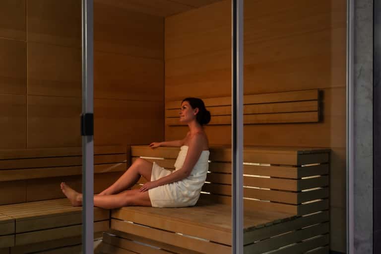 Woman relaxing in modern wooden sauna with horizontal slat paneling and glass doors