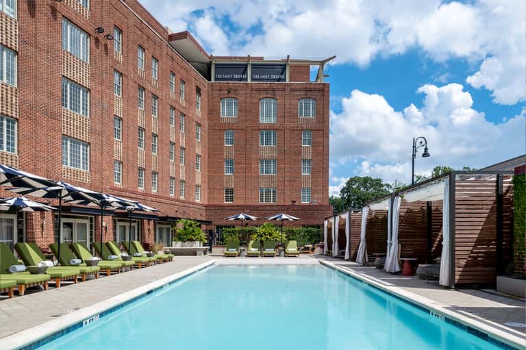 Resort pool with loungers, pergolas, brick building, and tropical landscaping in daylight