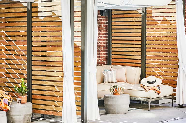Private outdoor lounge space with wooden slatted walls, cream seating, white curtains, and potted plant