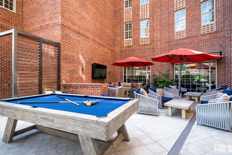 Courtyard patio with blue pool table, red umbrellas, white chairs, and brick building backdrop