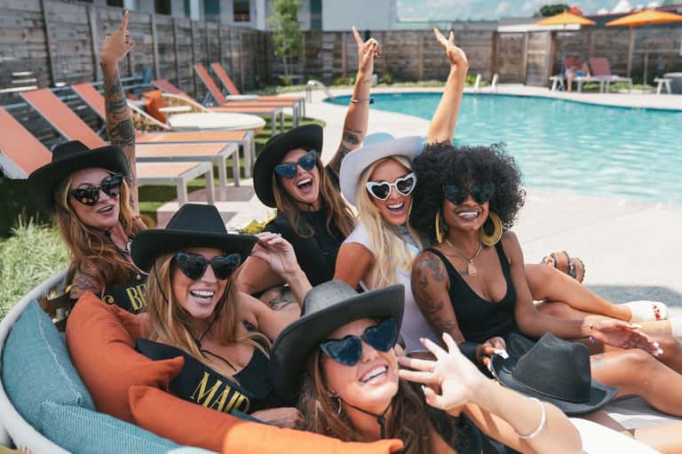 Group of six women in colorful outfits celebrating at a pool with loungers and umbrellas