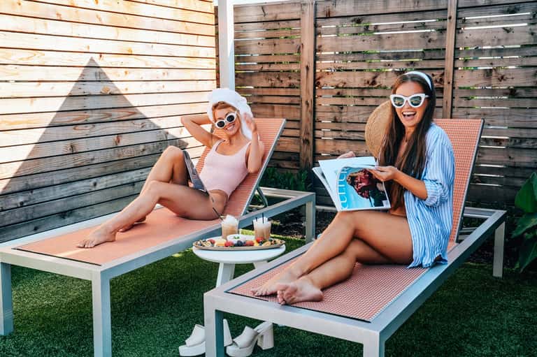 Two women relaxing on lounge chairs with food and drink, wooden privacy fence backdrop on manicured lawn