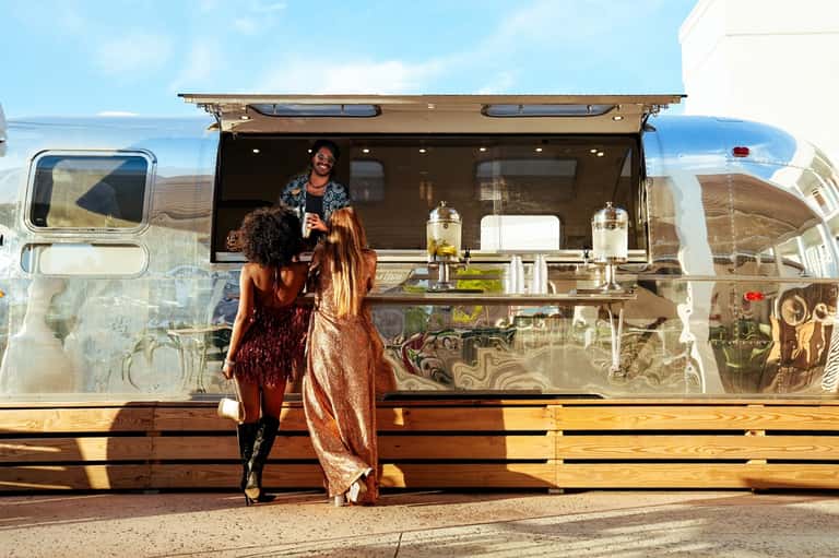 Women at vintage silver Airstream food truck service window on sunny day with beverages displayed