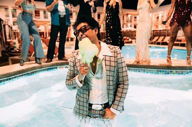 Man in plaid blazer enjoying cotton candy by pool with vintage resort setting and dressed guests