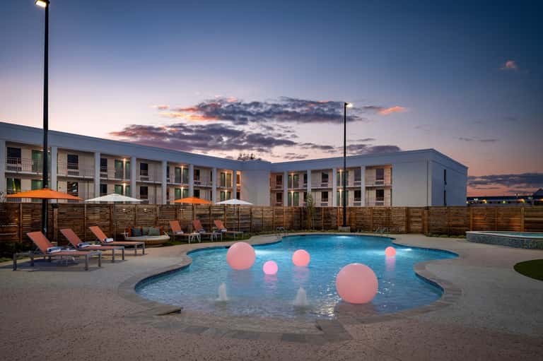 Resort pool at dusk with illuminated pink spheres, loungers, modern apartments, and wooden privacy fence
