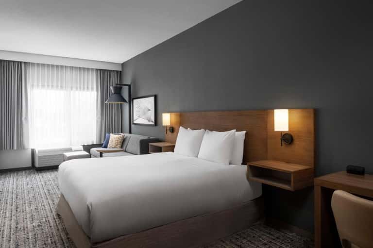 Contemporary hotel room with gray walls, wooden headboard with integrated lighting, white bedding, and seating area