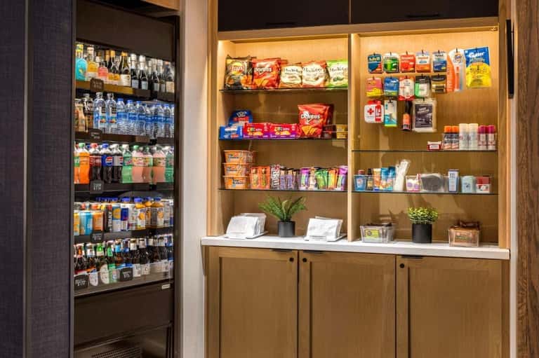 Hotel snack bar with refrigerated beverage case, wooden shelving with snacks, drinks, and toiletries