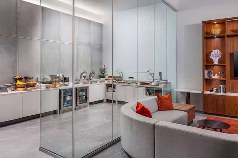 Contemporary hotel lounge with curved seating, kitchenette, glass partitions, and wood shelving with accent lighting