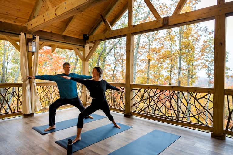 Couple practicing yoga on mats in a glass-enclosed pavilion overlooking autumn foliage