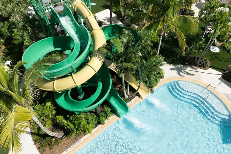 Aerial view of water slide in lush tropical resort grounds with palm trees and bright blue swimming pool