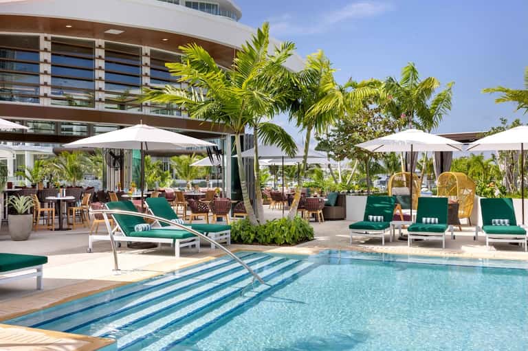 Resort pool area with green loungers, white umbrellas, palm trees, and modern waterfront building