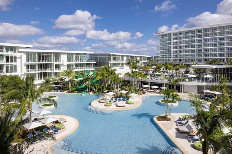Large resort pool complex with multiple pools, palm trees, lounge seating, and modern multi-story hotel buildings