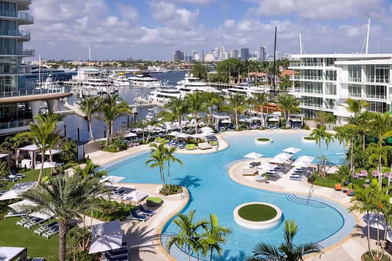 Waterfront resort pool with curved blue water, palm trees, loungers, marina with boats, and skyline in background