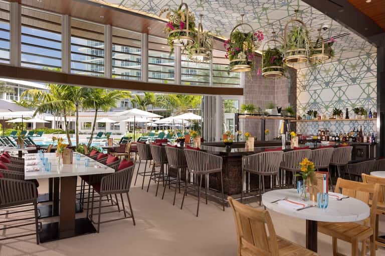 Modern restaurant bar with hanging floral installations, geometric ceiling, and outdoor pool view
