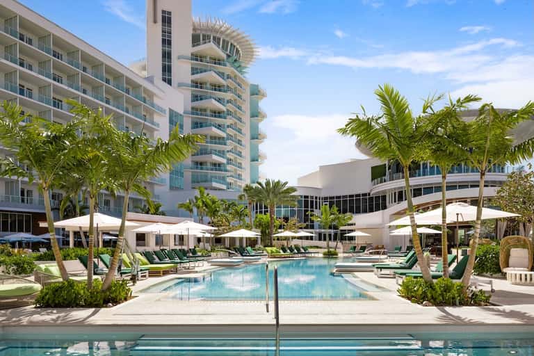 Resort pool surrounded by palm trees, lounge chairs, and modern hotel buildings