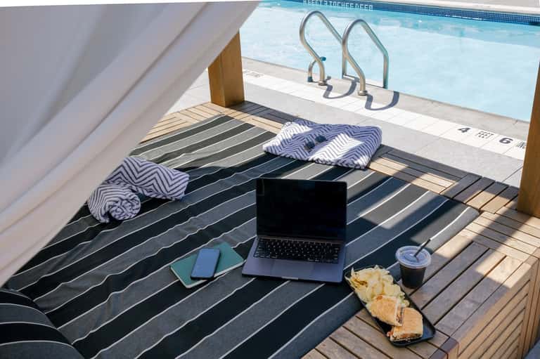 Poolside cabana with striped lounge bed, laptop, breakfast items, towels, and clear blue pool water visible
