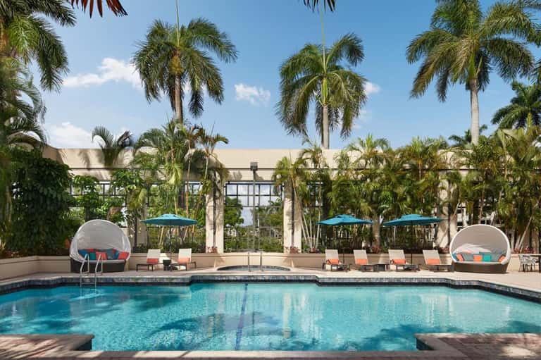 Resort pool with turquoise water, palm trees, lounge chairs, umbrellas, and modern architecture
