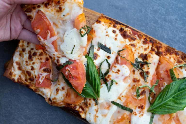 Slice of pizza with melted cheese, prosciutto, fresh basil leaves being held against blue background
