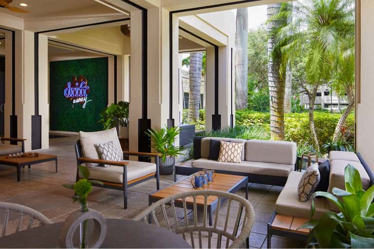 Covered outdoor lounge area with modern furniture, neon sign, and views of palm trees and grounds