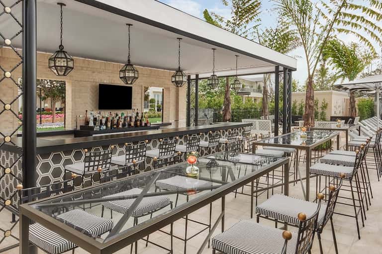 Upscale outdoor patio bar with black modern design, hanging pendant lights, geometric patterned chairs, and landscaping