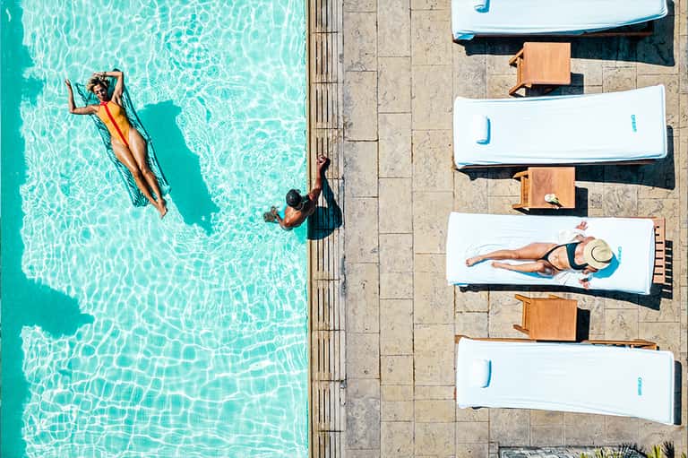 Aerial view of resort pool with swimmers, lounge beds with cushions, and concrete deck areas