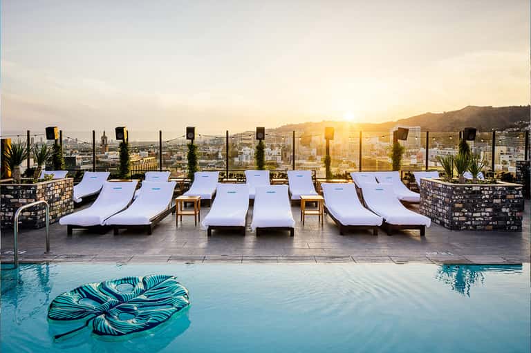 Luxury rooftop pool at sunset with white lounge chairs overlooking city skyline and mountains