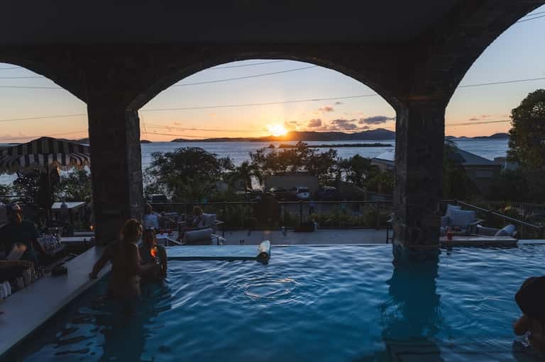 Infinity pool overlooking bay at sunset with concrete pillars and guests swimming