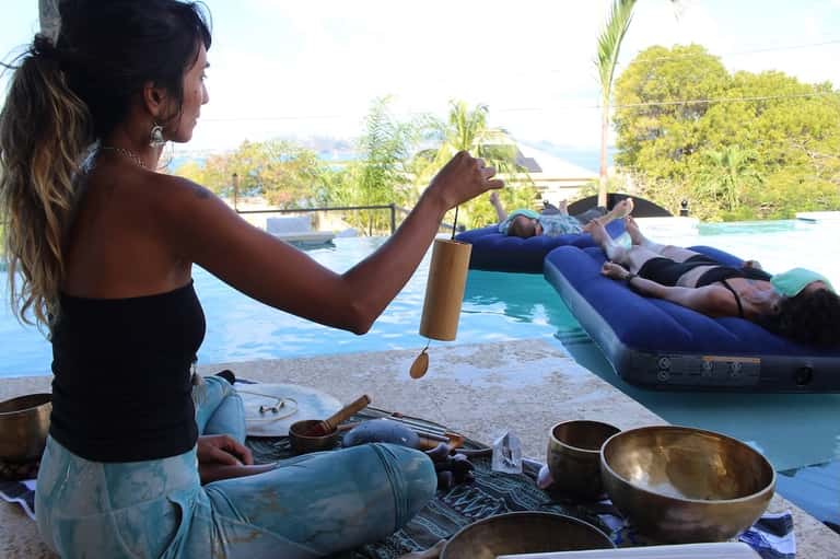 Woman performing sound bath meditation by pool with floating participants and tropical surroundings